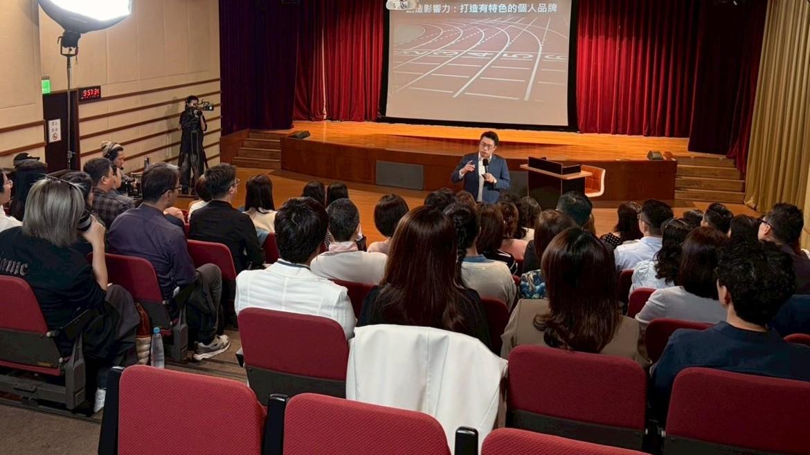 憲哥在台上演講，台下坐滿觀眾，旁邊攝影機與燈光同時在拍